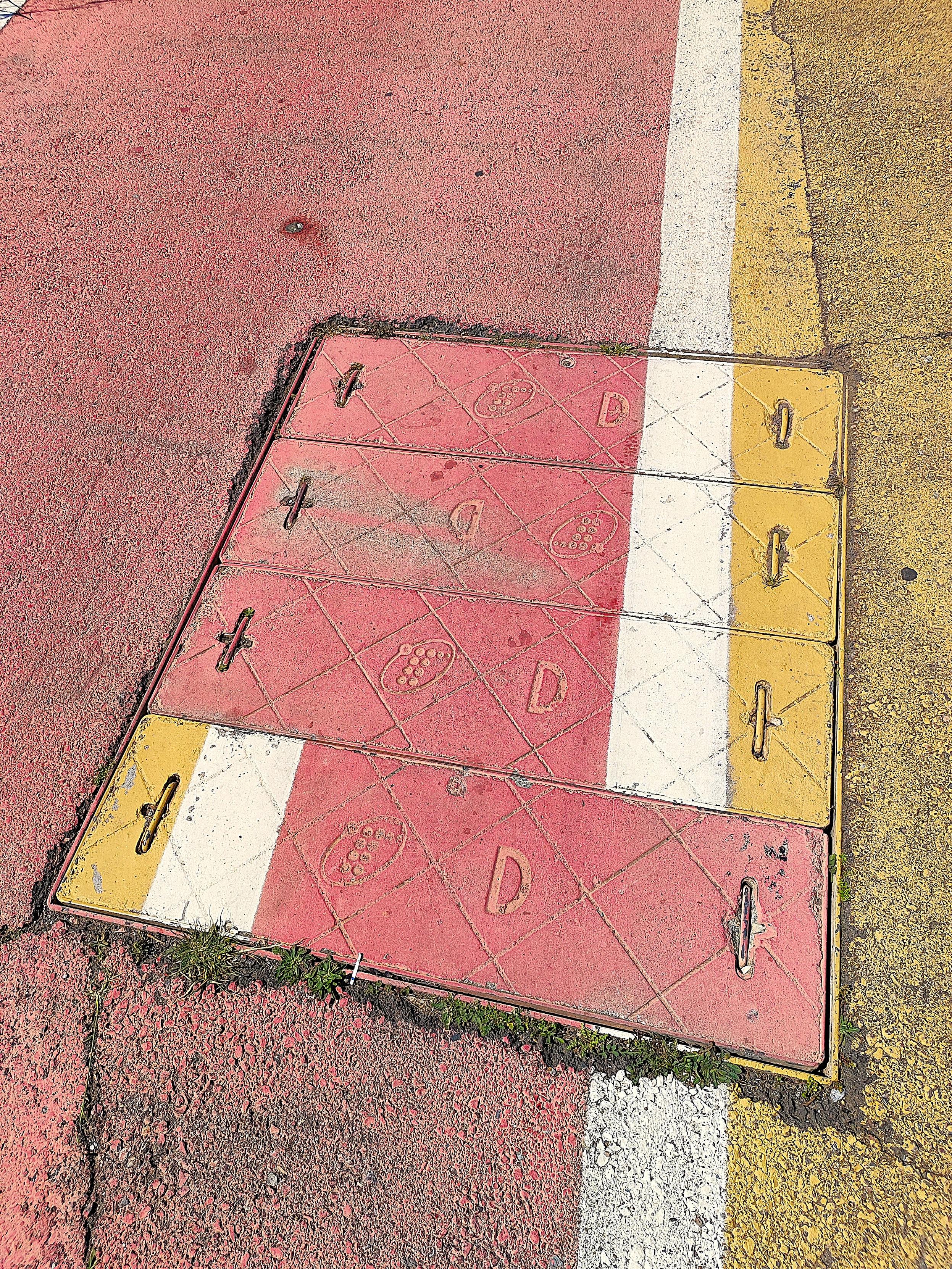 Vorera de color groc al costat d'un carril bici de color roig. Una franja de color blanc separa el carril bici de la vorera. Al centre de la imatge, en terra, una tapadora dividida en quatre segments. Tots els segments tenen gravats la lletra D i l'antic logo de Telefónica i estan pintats dels colors del carril bici, la franja blanca i la vorera. Tres dels segments estan col·locats de manera que els colors s'alineen amb la resta d'elements. Excepte el que està en primer pla, que està del revés i les franges groga i blanca queden en la zona del carril bici, que és roja.