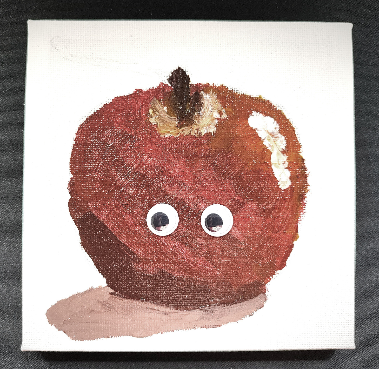 Llenç amb la imatge d'una poma roja pintada amb pintura acrílica. La poma té dos ulls de plàstic apegats.