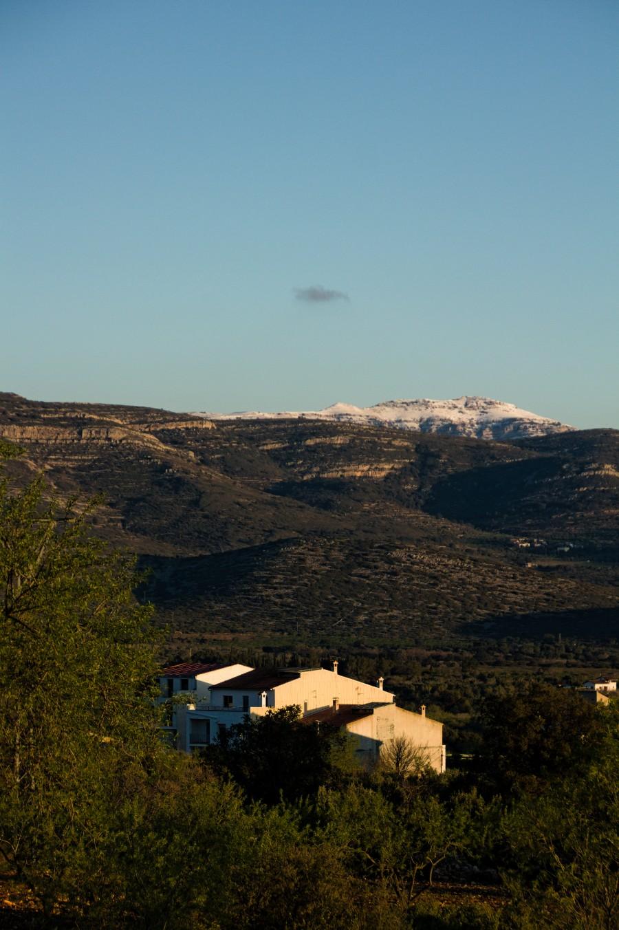 Paisatge rural amb algunes cases blanques envoltades de vegetació en primer pla i una muntanya amb cims nevats al fons. El cel és ras i blau.